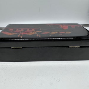 Puede incluir: Una caja de madera negra con tapa con bisagras. La tapa est&aacute; decorada con un dise&ntilde;o dorado y rojo de una escena con personas y &aacute;rboles.