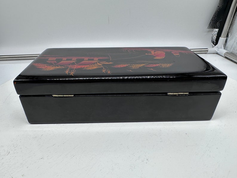 Puede incluir: Una caja de madera negra con tapa con bisagras. La tapa est&aacute; decorada con un dise&ntilde;o dorado y rojo de una escena con personas y &aacute;rboles.