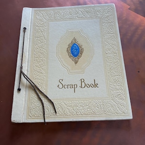 Puede incluir: Álbum de recortes crema con diseños florales en relieve y las palabras "Scrap Book" en una fuente cursiva. La portada presenta un elemento decorativo central azul y dorado. La encuadernación es de cordón.