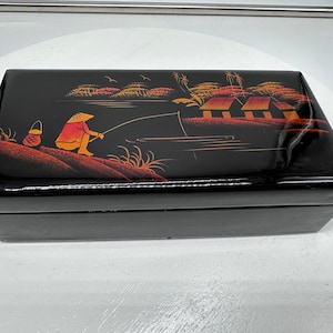 Puede incluir: Caja de madera lacada en negro con una escena pintada a mano de una persona pescando en un r&iacute;o con casas en el fondo. La escena est&aacute; pintada en tonos de naranja, rojo y amarillo.