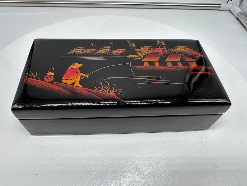 Puede incluir: Caja de madera lacada en negro con una escena pintada a mano de una persona pescando en un r&iacute;o con casas en el fondo. La escena est&aacute; pintada en tonos de naranja, rojo y amarillo.