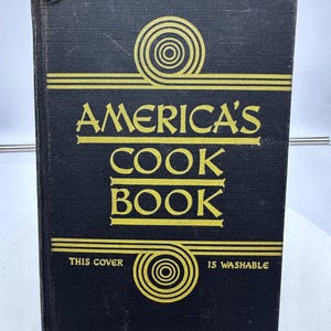 May include: A black cookbook with gold lettering that reads "America's Cook Book". The cover has a gold design with concentric circles and the text "This cover is washable".
