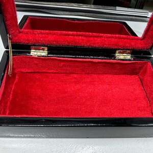 Puede incluir: Caja de madera negra con interior de terciopelo rojo y tapa con bisagras. La caja tiene un cierre y bisagras doradas.