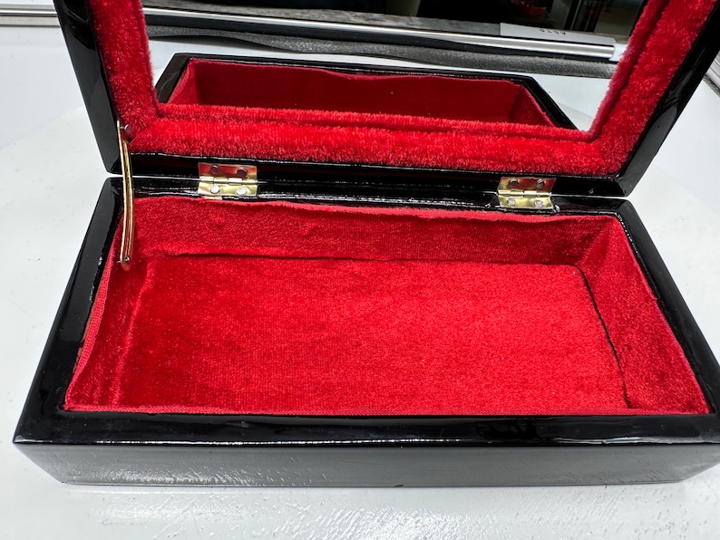 Puede incluir: Caja de madera negra con interior de terciopelo rojo y tapa con bisagras. La caja tiene un cierre y bisagras doradas.
