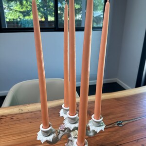 May include: A white ceramic candelabra with five arms, each holding a peach-coloured candle. The candelabra is decorated with floral details and has a delicate, ornate design.