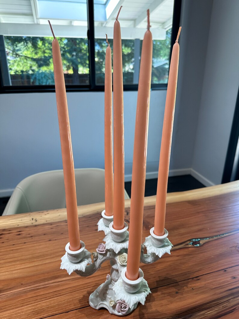 May include: A white ceramic candelabra with five arms, each holding a peach-coloured candle. The candelabra is decorated with floral details and has a delicate, ornate design.