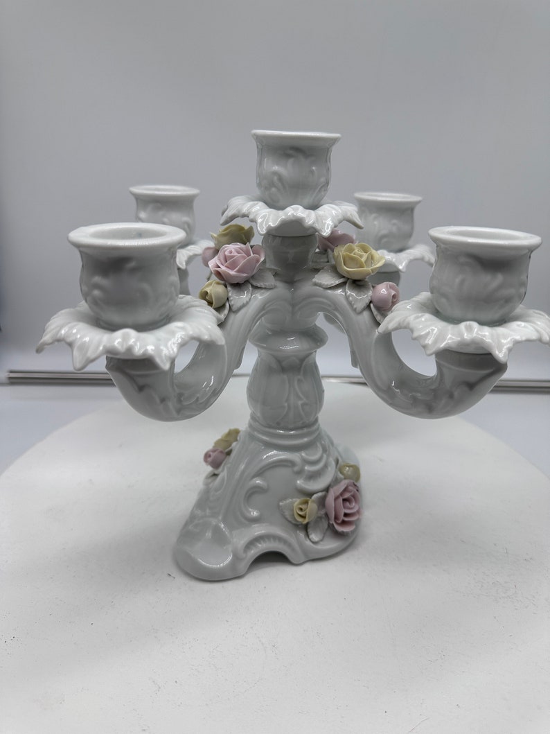 May include: White porcelain candelabra with five candle holders and floral accents. The candelabra is decorated with delicate pink and yellow roses.