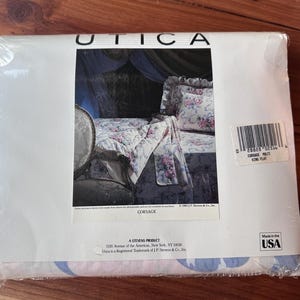 Könnte beinhalten: Versiegeltes Paket mit Bettwäsche der Marke Utica. Die Verpackung zeigt ein Bett mit floral gemusterten Bettlaken und Kissenbezügen. Der Text "CORSAGE" ist sichtbar, zusammen mit "Made in the USA."