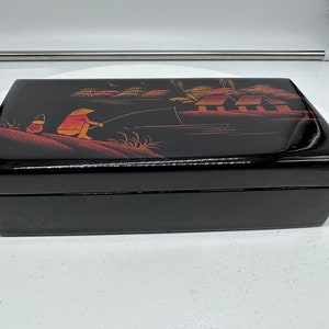 Puede incluir: Caja de madera negra con una escena pintada de una persona pescando en un bote con casas en el fondo. La escena est&aacute; pintada en rojo, naranja y amarillo.