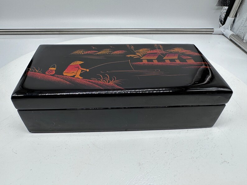 Puede incluir: Caja de madera negra con una escena pintada de una persona pescando en un bote con casas en el fondo. La escena est&aacute; pintada en rojo, naranja y amarillo.