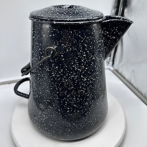 May include: Black enamel camping coffee pot with white speckles and a wire handle. The pot has a spout and a lid.