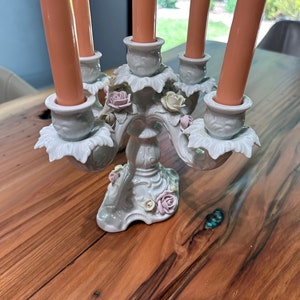 May include: A white porcelain candelabra with five candle holders and floral accents. The candelabra is decorated with delicate pink and white flowers and leaves. It is sitting on a wooden table.