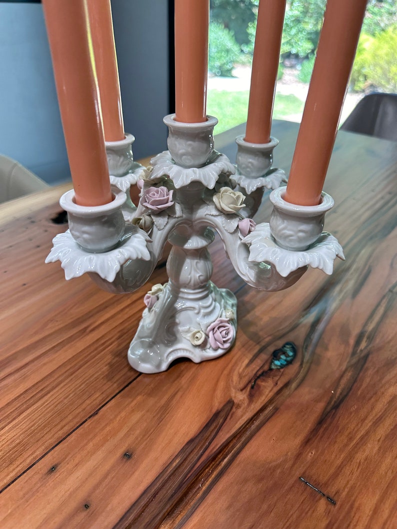 May include: A white porcelain candelabra with five candle holders and floral accents. The candelabra is decorated with delicate pink and white flowers and leaves. It is sitting on a wooden table.