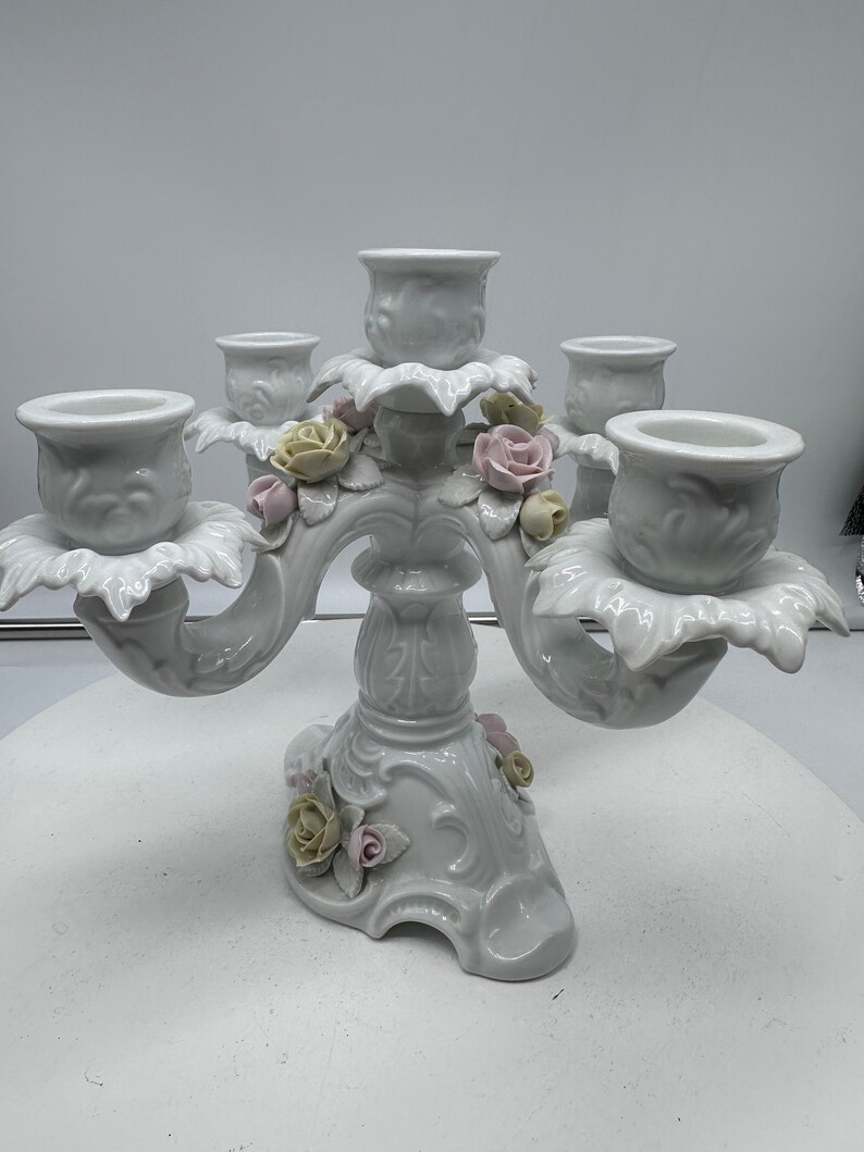 May include: A white porcelain five-arm candelabra with floral accents. Each arm features a delicate rose and leaf design. The base is intricately detailed with a floral motif.