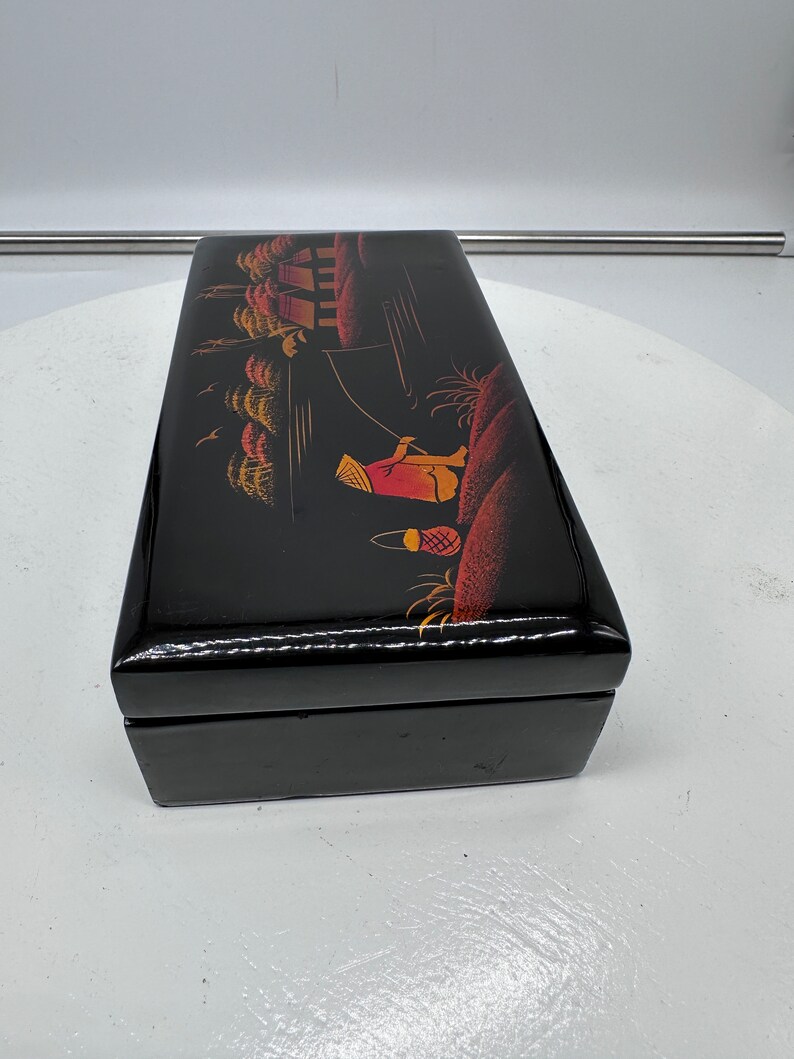 Puede incluir: Caja de madera negra con una escena pintada a mano de una persona con un sombrero y ropa tradicionales pescando en un r&iacute;o con casas y &aacute;rboles en el fondo.