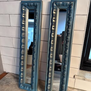 Puede incluir: Dos espejos de pared rectangulares con marcos azul turquesa ornamentados. Los marcos tienen un patrón texturizado y un borde interior dorado. Los espejos están apoyados contra una pared de azulejos blancos y una encimera.