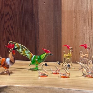 May include: A display of glass animal ornaments, featuring a rooster, a pheasant, swans, and deer. The rooster is brown, orange, and white. The pheasant is green and yellow. The swans are clear with orange bases.