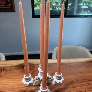 May include: A white ceramic candelabra with five arms, each holding a peach-coloured candle. The candelabra is decorated with floral details and sits on a wooden table.