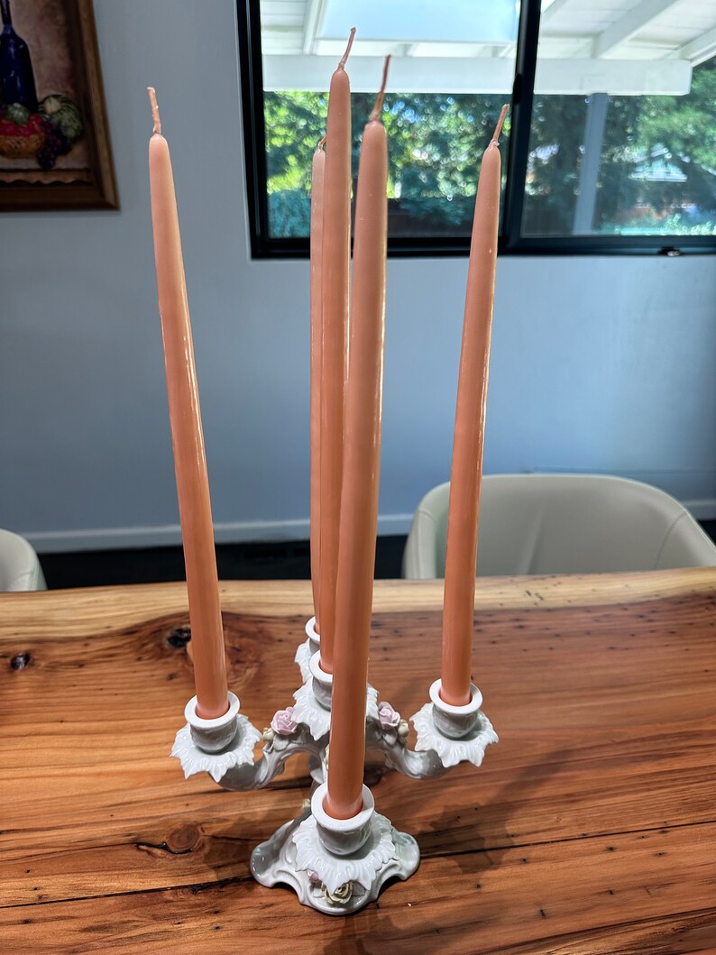 May include: A white ceramic candelabra with five arms, each holding a peach-coloured candle. The candelabra is decorated with floral details and sits on a wooden table.
