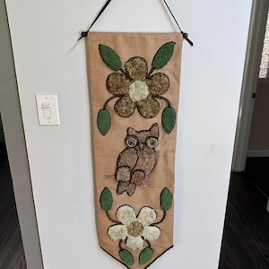 以下が含まれることがあります： 茶色のジュート製の壁掛けで、茶色のフクロウと緑と茶色の花が2つ付いています。フクロウは壁掛けの中央にあり、花は上下に配置されています。壁掛けの下部には茶色のタッセルが付いています。