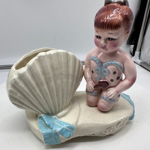 Puede incluir: Un macetero de cerámica con forma de concha de mar con una figura de niña sentada encima. La niña lleva un traje de baño rosa con lunares azules y un lazo azul en el pelo. El macetero es blanco con detalles azules.