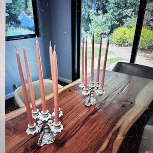 May include: Two ornate white candle holders with a floral design, each holding six tall, slender, peach-coloured candles on a wooden table.