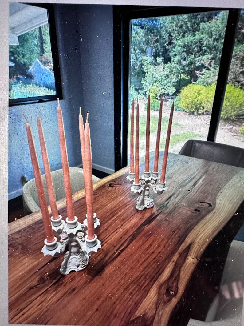 May include: Two ornate white candle holders with a floral design, each holding six tall, slender, peach-coloured candles on a wooden table.