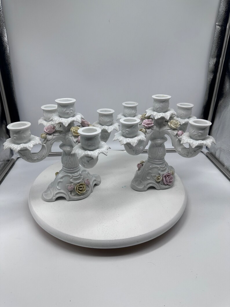 May include: A pair of white porcelain candle holders with floral accents. Each holder has five candle cups and features delicate pink and yellow roses.