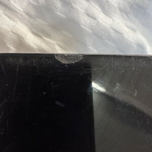 Puede incluir: Una superficie de vidrio negro con un peque&ntilde;o astillado en el borde superior. La superficie est&aacute; rayada y muestra signos de desgaste.