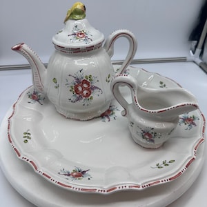 Puede incluir: Un juego de té de cerámica blanca con una tetera, una jarra de crema y un plato decorativo. La tetera tiene un adorno de pájaro y diseños florales. La jarra de crema y el plato también tienen diseños florales.