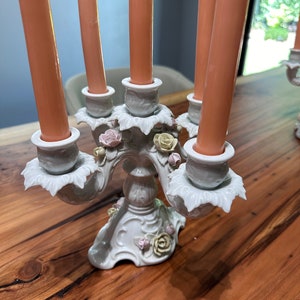 May include: A white porcelain candelabra with five arms, each featuring a delicate floral design and a pink candle. The base of the candelabra is decorated with roses and leaves.