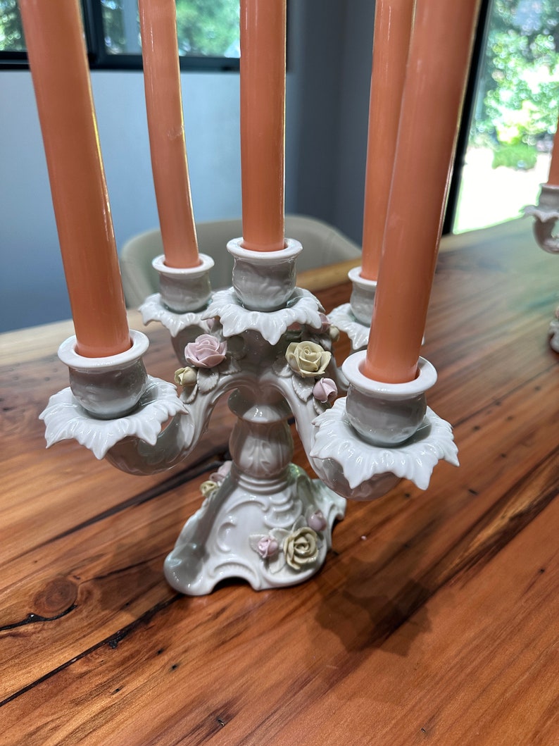 May include: A white porcelain candelabra with five arms, each featuring a delicate floral design and a pink candle. The base of the candelabra is decorated with roses and leaves.