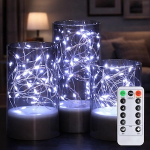 Op de afbeelding: Drie glazen cilindervazen met witte LED-snoerlampjes. De vazen hebben een zilveren basis en verschillende hoogtes. Een afstandsbediening met "ON", "OFF" knoppen en nummers is zichtbaar.