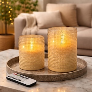 May include: Two cylindrical, textured, amber-colored LED candles of different sizes sit on a round tray. A remote control is in the foreground. The candles are lit, creating a warm glow. The background includes a sofa and a small tree.