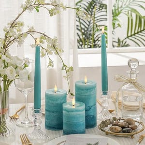 May include: A dining table setting with teal candles of different sizes, some with lit flames, and clear glass candle holders. White flowers and foliage surround the candles. A decorative glass bottle and a plate with a floral pattern are also visible.