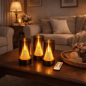May include: Three LED candle lamps with a warm glow. Each lamp has a gold-coloured metal base and a clear glass cylinder. The lamps are on a wooden table. A remote control is visible on the table.
