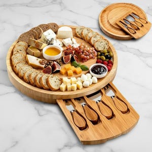 May include: A round bamboo cheese board with a variety of cheeses, bread, fruit, and condiments. The board includes a pull-out drawer with cheese knives and a smaller serving plate. The arrangement is on a white marble surface.