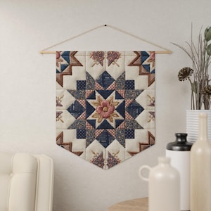 Puede incluir: Un tapiz de pared acolchado con un diseño de estrella. La estrella central presenta un motivo floral rosa, rodeado de patrones geométricos azules y crema. El tapiz está suspendido de una barra de madera y cuerda.