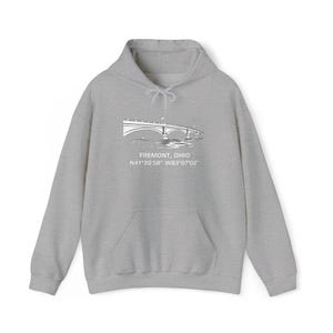 Puede incluir: Una sudadera con capucha gris claro con un bolsillo delantero y una capucha con cordón. La parte delantera presenta un gráfico blanco de un puente con el texto "FREMONT, OHIO" y coordenadas. Una sudadera informal y cómoda.