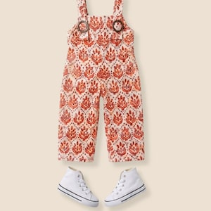 May include: A sleeveless jumpsuit featuring a repeating orange and red floral pattern on a cream background, with two circular brown buckles at the shoulder straps, paired with a set of white high-top canvas sneakers with black trim.