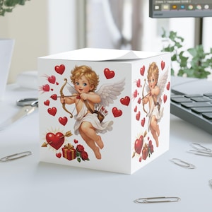 Könnte beinhalten: Weißer, würfelförmiger Notizblock mit einem Cartoon-Cupido-Motiv, komplett mit Pfeil und Bogen und roten Herzen. Der Cupido hat blondes Haar, weiße Flügel und eine weiße Toga. Eine Geschenkbox und Rosen sind ebenfalls abgebildet.