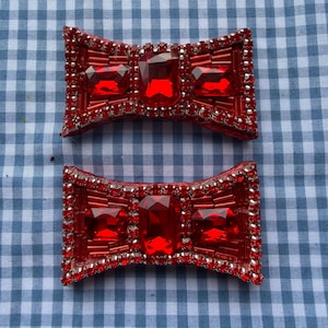 May include: Two red bow-shaped hair clips with sparkling red gems and silver accents. The bows have a central rectangular gem and smaller gems around the edges. They are set against a blue and white checkered background.