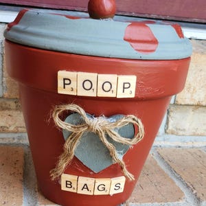 Puede incluir: Una maceta de color terracota con tapa, pintada con detalles en gris y rojo. La maceta tiene las palabras "POOP" y "BAGS" escritas con fichas de Scrabble. Un lazo de cuerda y una decoración en forma de corazón están adheridos al frente.