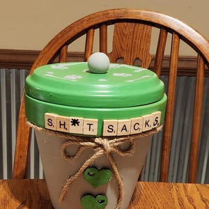 May include: A hand-painted, decorative flower pot with a green lid and a white knob. The pot is gray with a jute bow and two green heart accents. The lid has white paw prints and the word "SHT*SACKS" spelled out with Scrabble tiles.