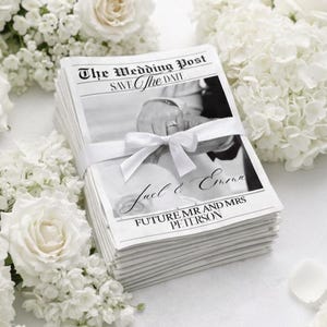 May include: A pile of wedding announcement cards, resembling newspapers, tied with a white ribbon. The cards feature the headline "The Wedding Post", names "Jael & Emma", and "FUTURE MR AND MRS PETERSON". White flowers, including roses and hydrangeas, surround the cards.