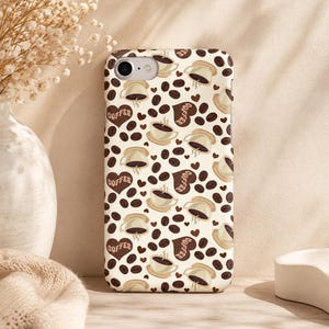 May include: A phone case with a repeating pattern of coffee cups, coffee beans, and heart-shaped coffee bean designs. The background is off-white. The coffee cups are beige with brown coffee and a brown handle. The heart-shaped designs have the word "COFFEE".