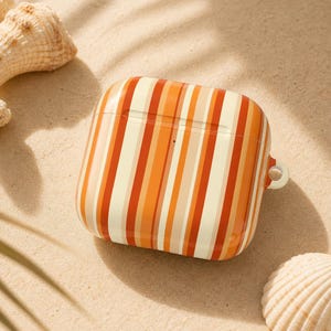May include: A square, striped AirPod case in shades of orange, cream, and tan. The case is resting on a bed of sand, with seashells in the background. The case has a small loop for attaching to a keychain.