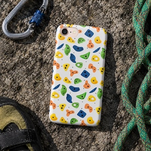 May include: White phone case with a colorful climbing hold pattern in yellow, green, blue, and orange. The case is on a rock surface with climbing gear, including a carabiner and rope, in the background.