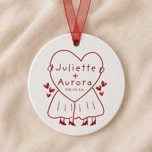 May include: A round white ceramic ornament featuring a red line drawing of two figures holding a large heart. The heart contains the text Juliette + Aurora and the date 08.19.26. Small red hearts surround the figures, and a red ribbon is attached.