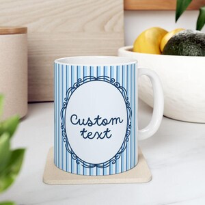 Può includere: Tazza in ceramica bianca con motivo a righe blu e bianche. La tazza presenta un design ovale con le parole "Custom text" in corsivo. La tazza è appoggiata su un sottobicchiere beige.